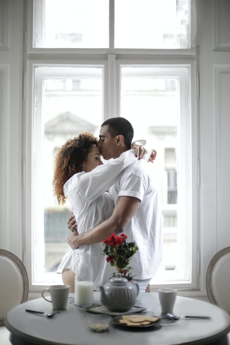 Couple Hugging And Kissing While Standing Near Window At Home