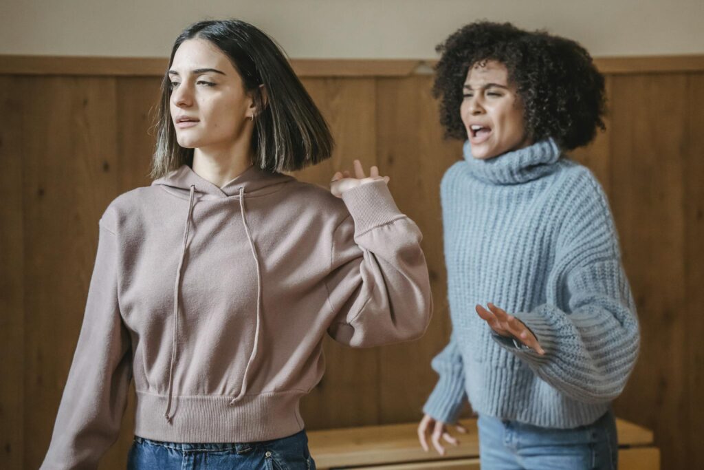 Mad African American female in warm sweater screaming at irritated female while having argument in light room with wooden walls