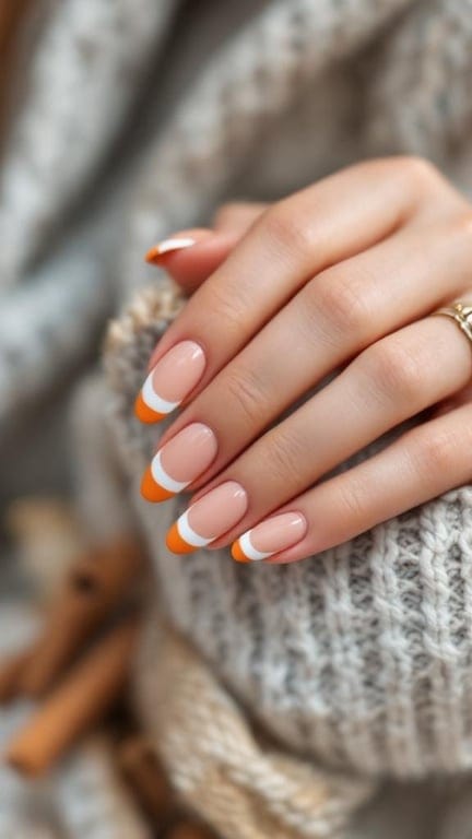 A close-up of hands with pumpkin spice French tip nails, featuring a nude base and orange tips, resting on a cozy knitted sweater.