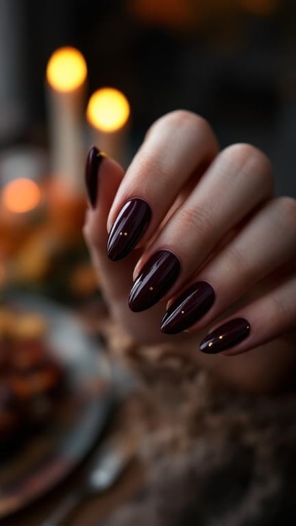 A hand with deep plum nails featuring gold dot cuticles, set against a cozy background.