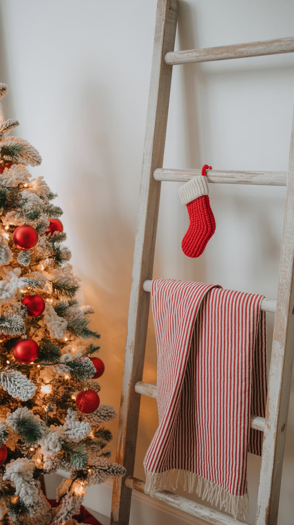 A Christmas tree with red ornaments next to a farmhouse ladder holding a red stocking and a striped blanket.