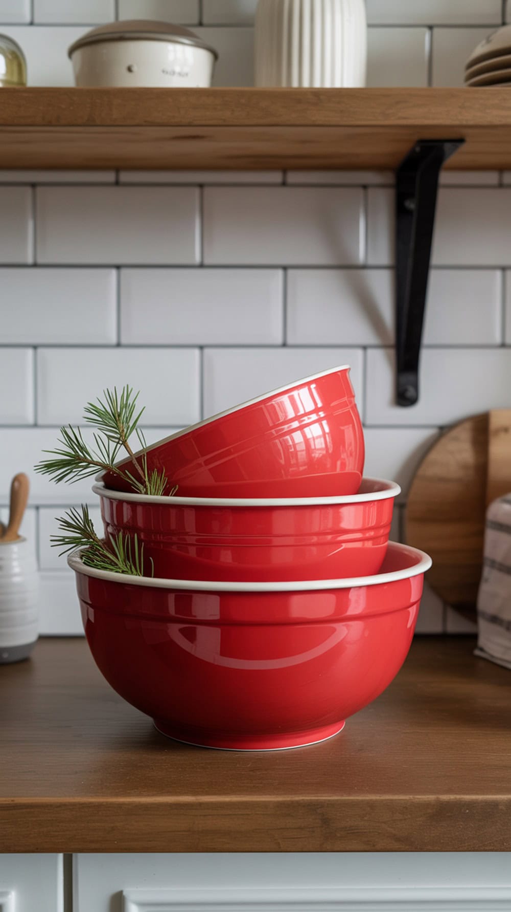 A stack of red mixing bowls with pine sprigs on a wooden shelf in a modern farmhouse kitchen.