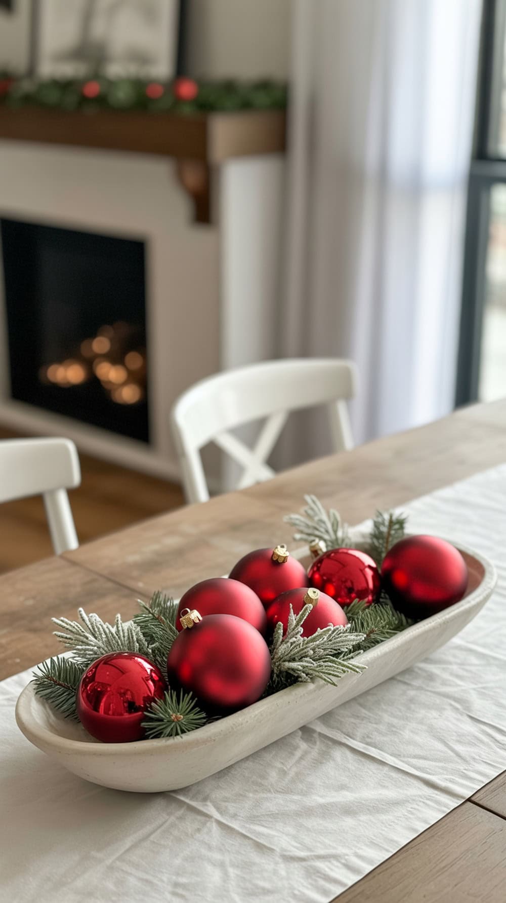 A white dough bowl filled with red ornaments and greenery on a wooden table.