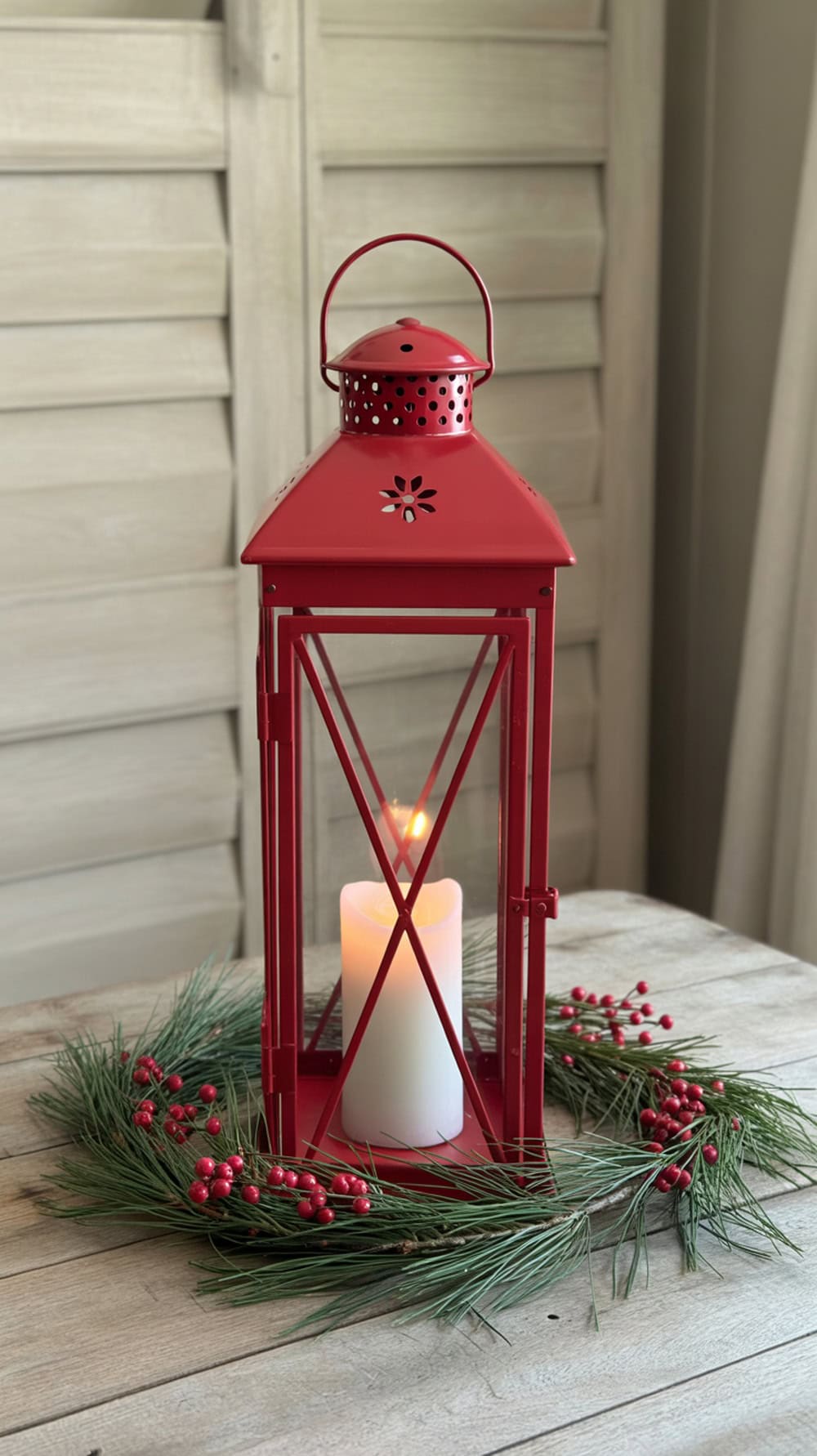 A red metal lantern with a candle inside, surrounded by a pine wreath with red berries, on a wooden table.