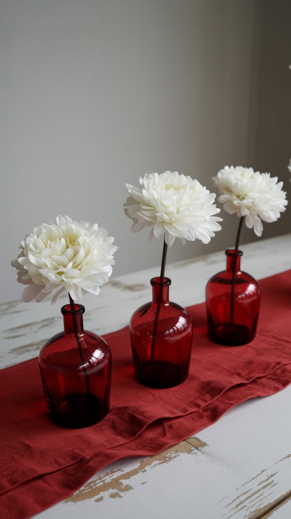 Three red glass vases with white flowers on a red table runner.