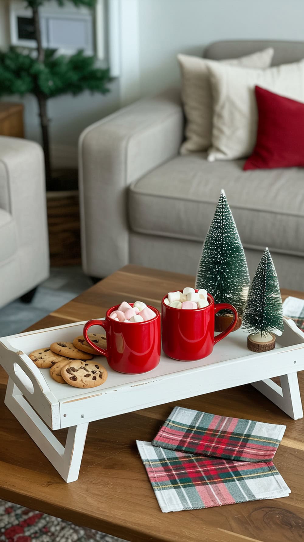 A white tray with red mugs filled with marshmallows, cookies, and small evergreen trees on a wooden table.