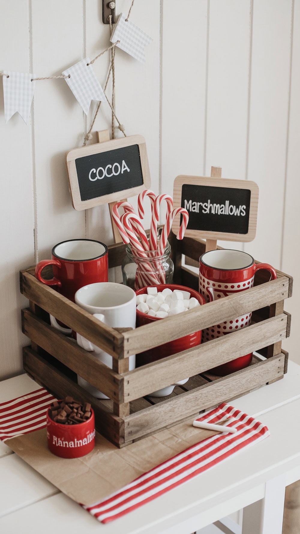 A mini hot cocoa bar with red mugs, candy canes, marshmallows, and a wooden crate setup.