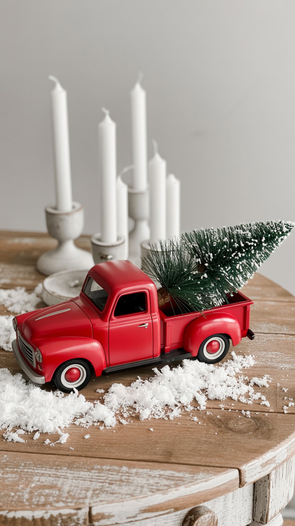 A mini red truck with a tree figurine on a wooden table with white candles and faux snow.