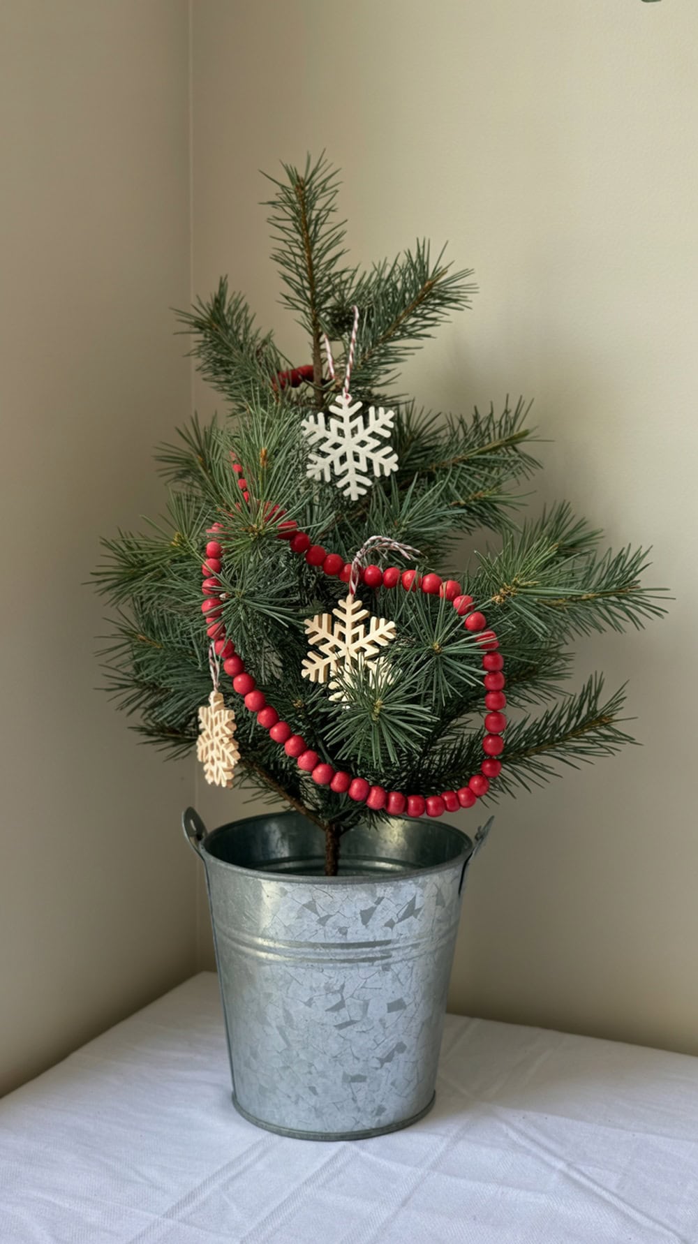 A mini tabletop Christmas tree with red beads and wooden snowflake ornaments in a galvanized bucket.