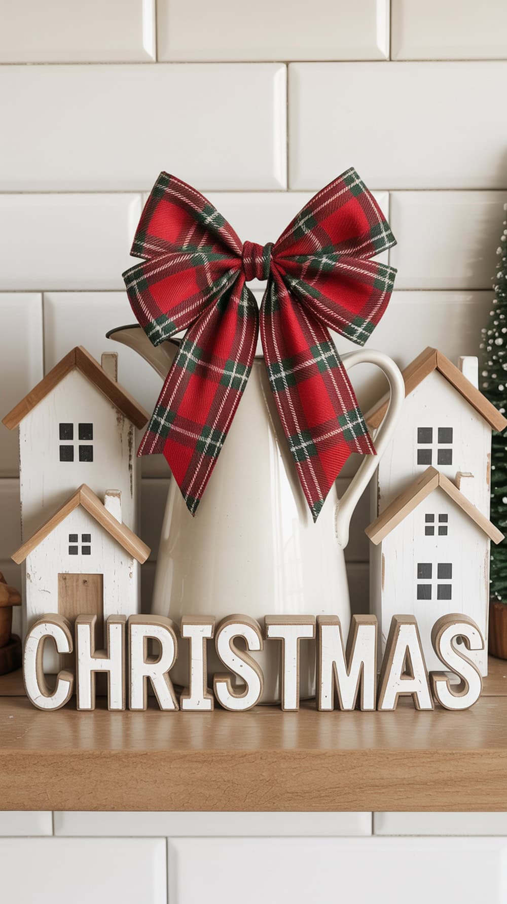 A white pitcher adorned with a red plaid bow, surrounded by white decorative houses on a kitchen shelf.