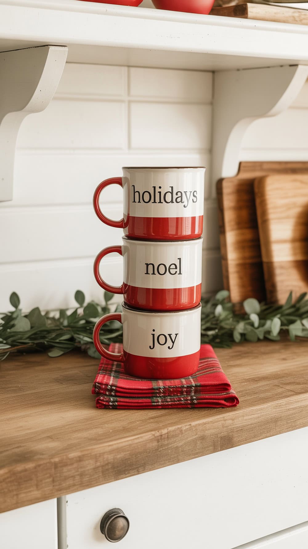 A shelf displaying stacked red and white mugs with holiday phrases, surrounded by greenery and a plaid napkin.