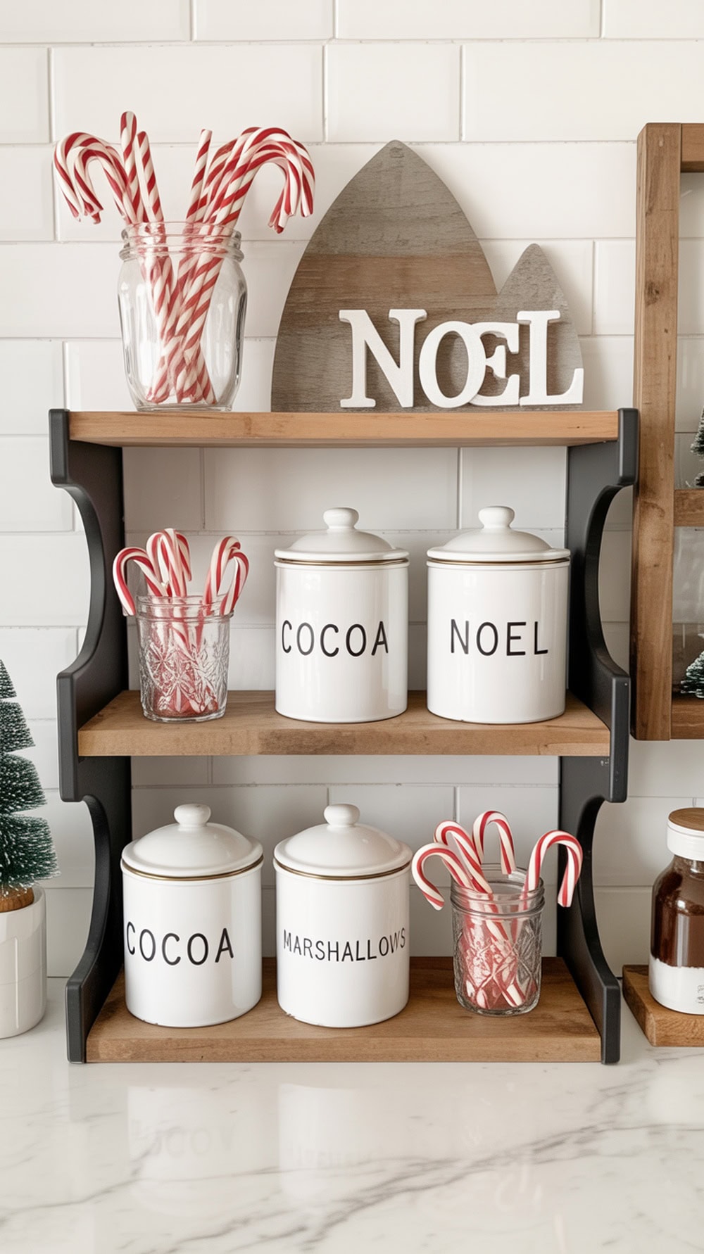 A cozy hot cocoa station shelf with white canisters, glass jars filled with candy canes, and decorative trees.