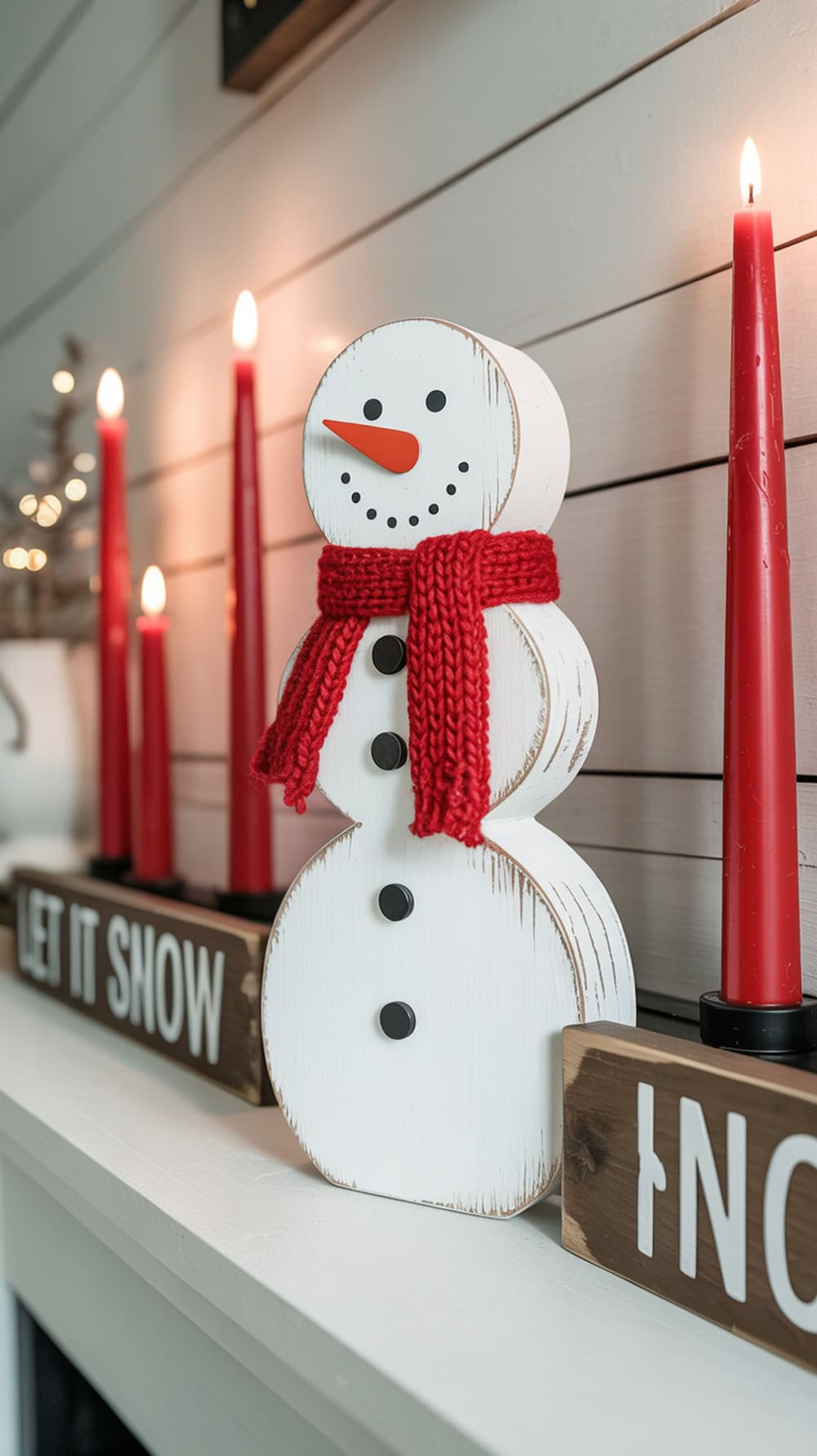 A wooden snowman decoration with a red scarf, surrounded by red candles and holiday signs on a shelf.