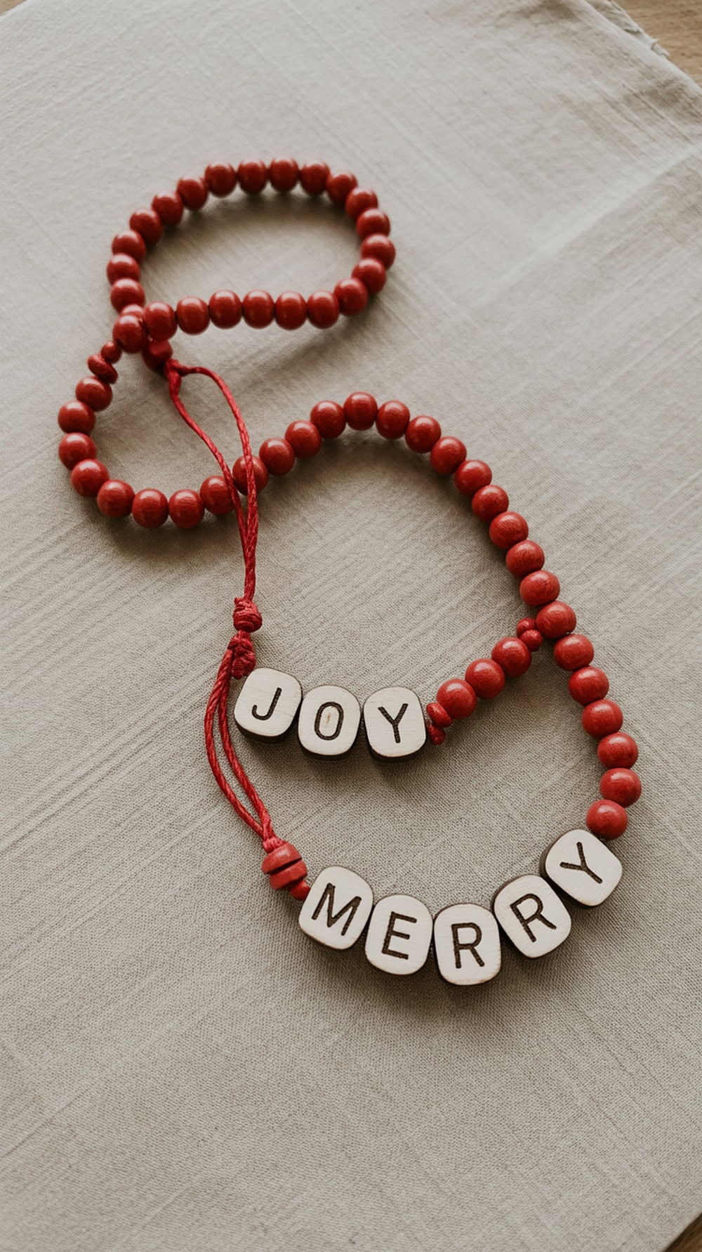 Red wooden beads with white tags spelling 'Joy' and 'Merry' on a neutral fabric background