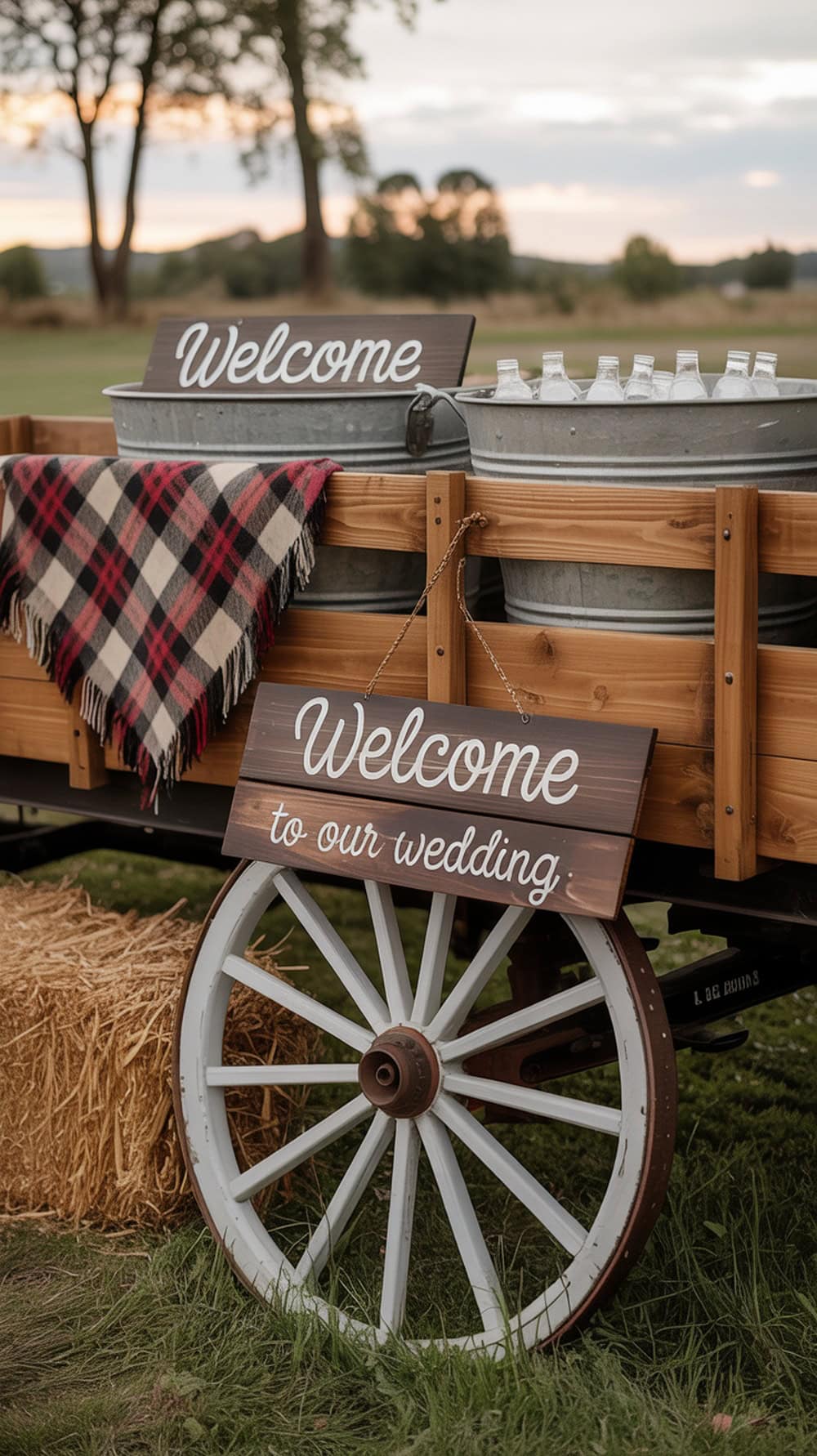 A rustic wagon display with a welcome sign, filled with drinks and a plaid blanket, set in a country setting.