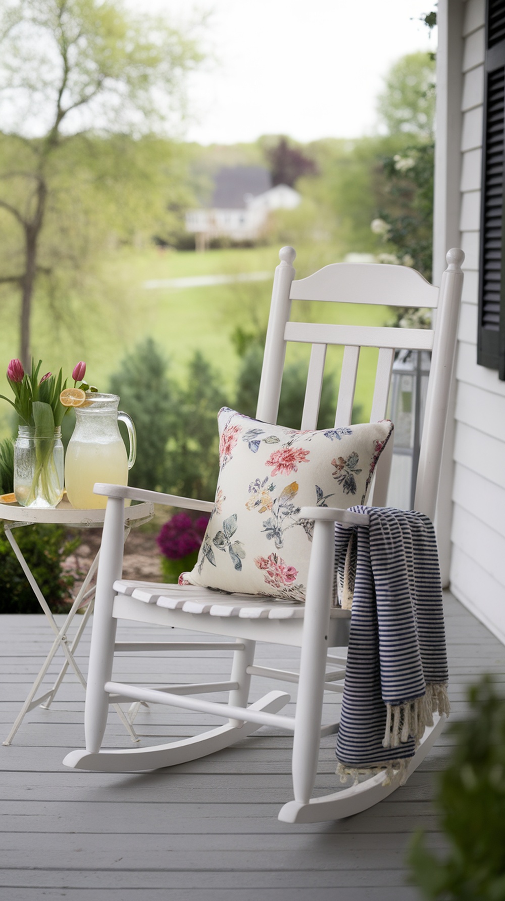 A cozy white rocking chair on a porch with a floral pillow, a small table with lemonade, and tulips in a jar.