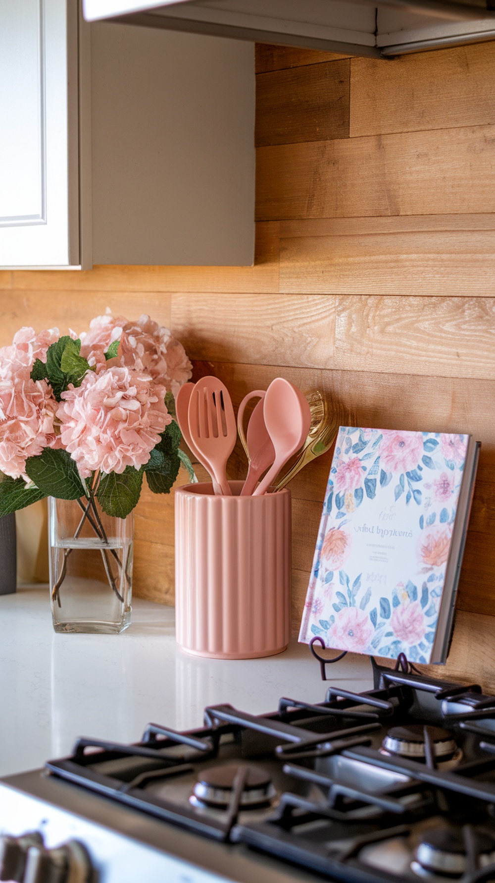 A kitchen counter styled with blush and white decor, featuring pink flowers, utensils, and a floral recipe book.