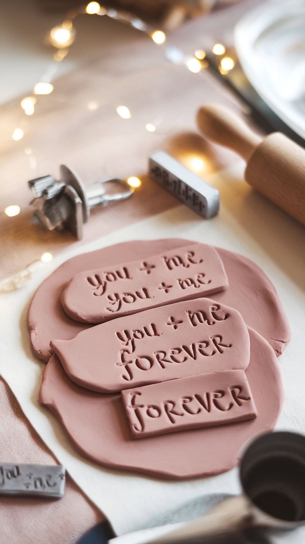 Clay pieces with stamped messages on a soft surface, surrounded by crafting tools and fairy lights.