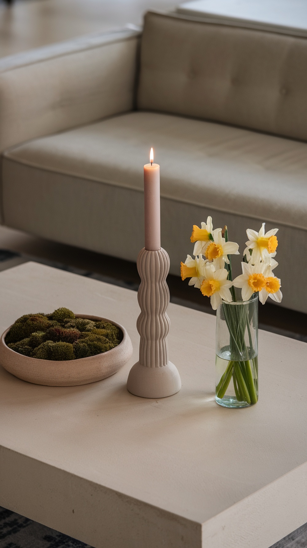 A modern coffee table styled with a pastel candle, a vase of daffodils, and a bowl of moss.