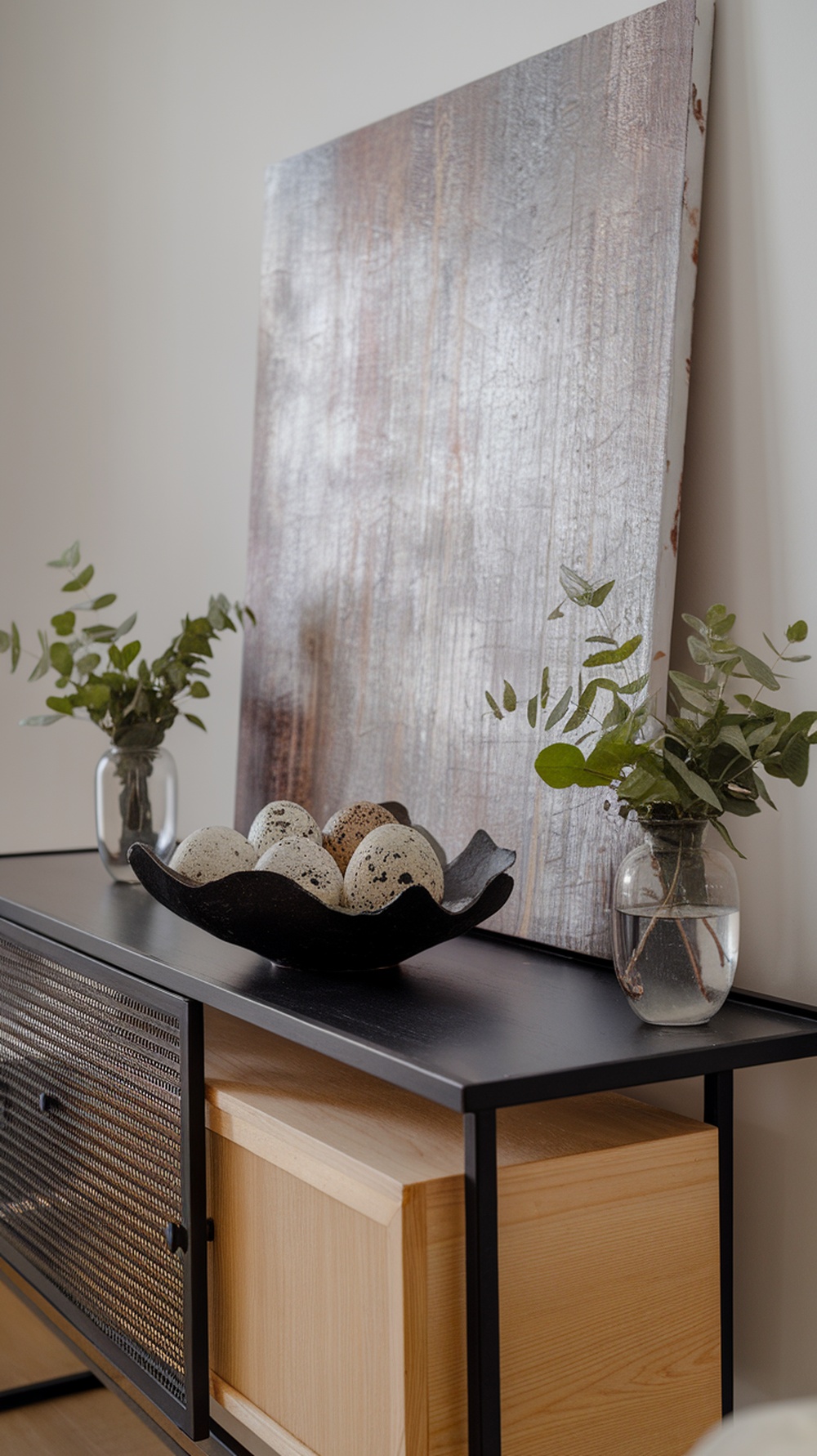 A modern entry console with abstract art, decorative eggs, and fresh greenery.