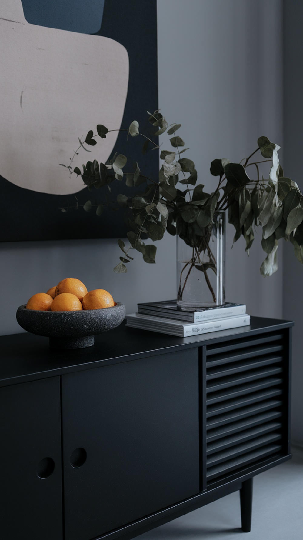 A modern black sideboard with a bowl of oranges and a vase of eucalyptus on top, alongside stacked books.