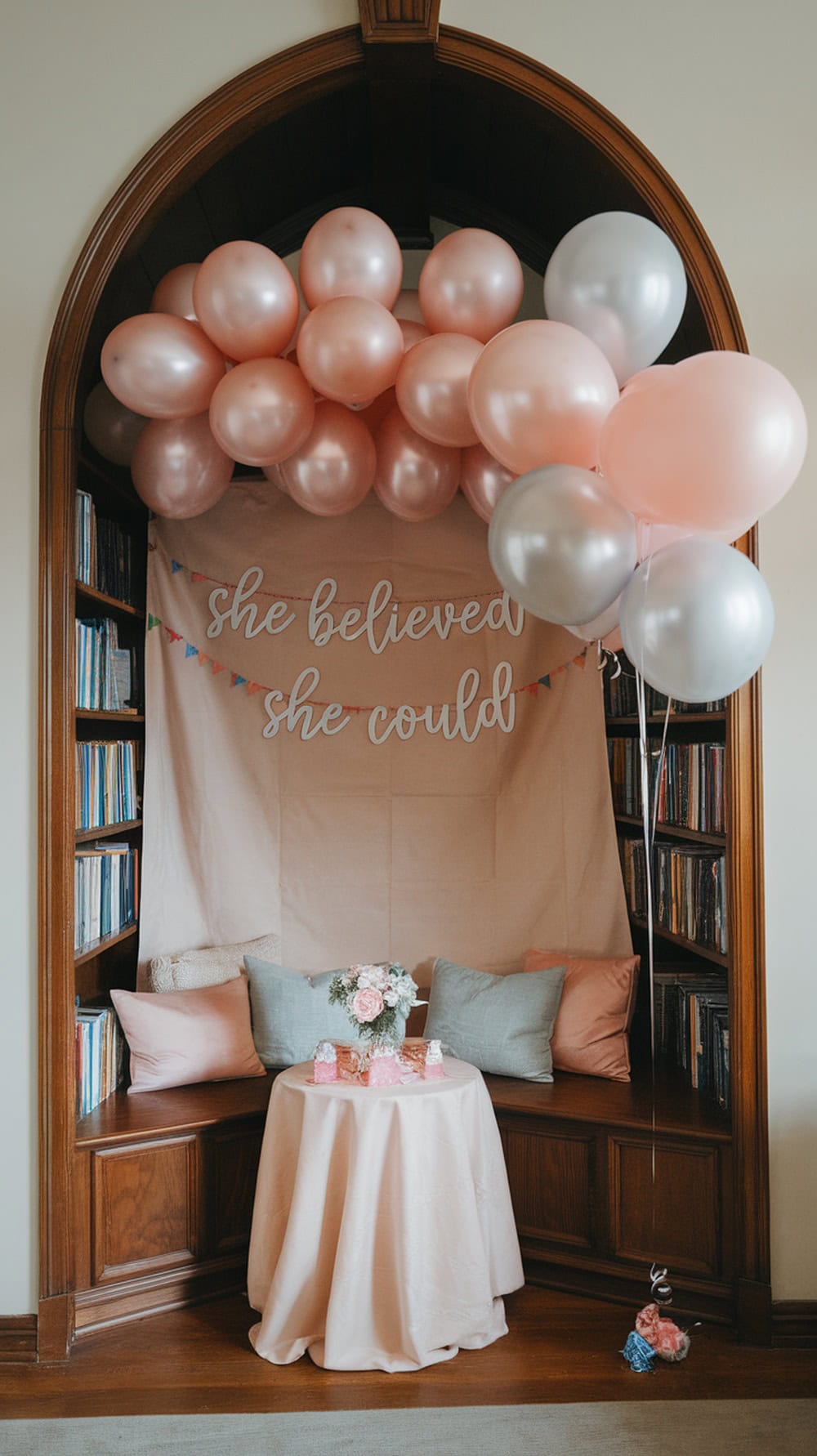A cozy photo corner for a graduation party with pink and silver balloons, a soft pink backdrop, and a small table with flowers and treats.