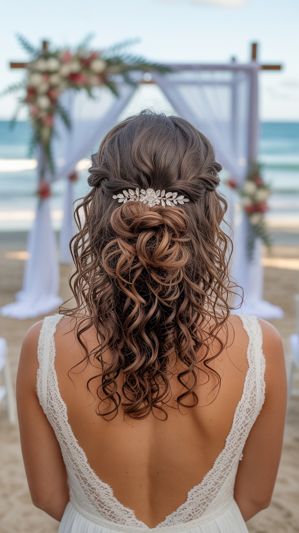 A woman with curly hair styled in a half up knot with a crystal barrette, standing at a beach wedding.