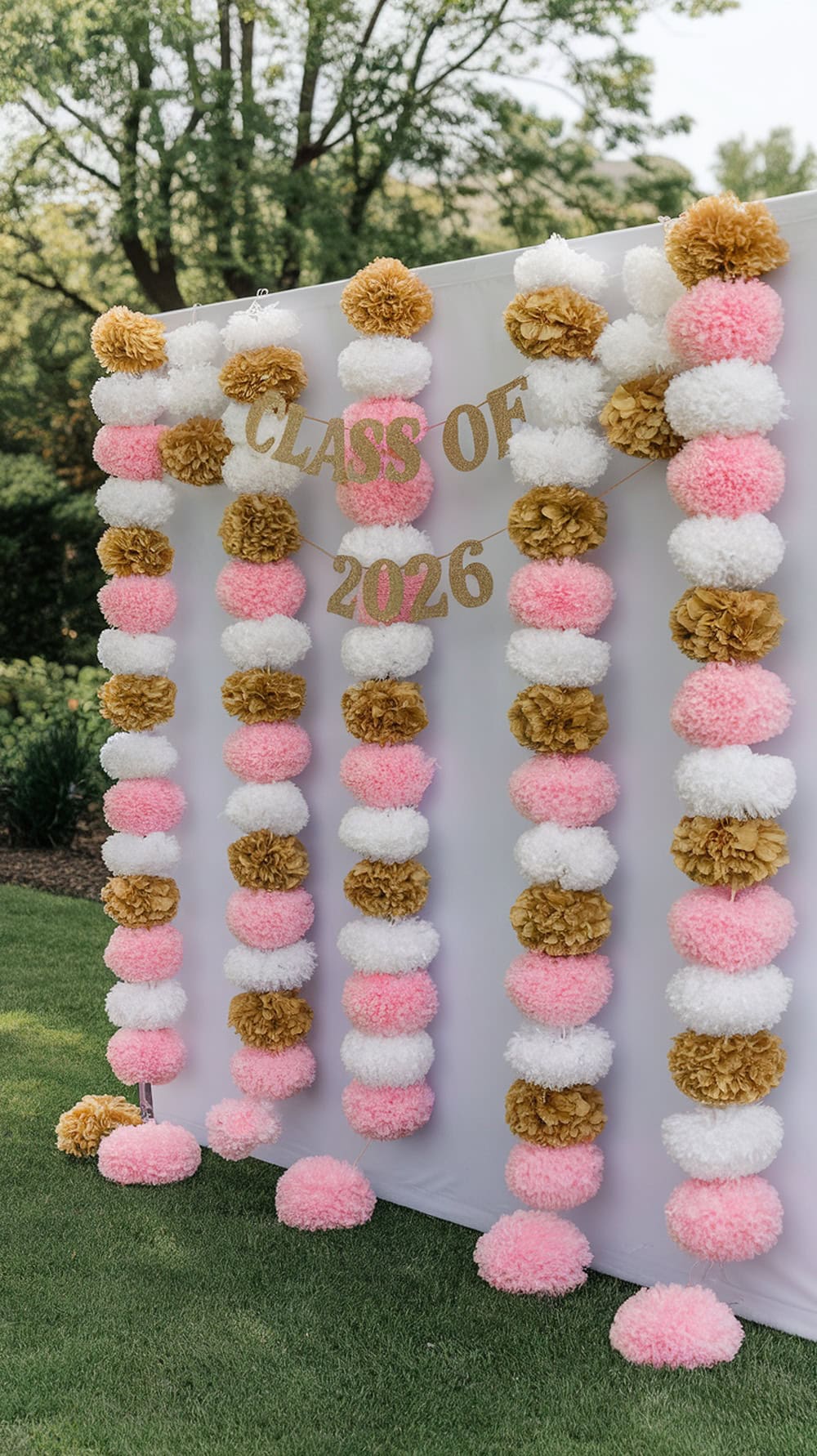 A colorful pom pom garland backdrop for a graduation party, featuring pink, white, and gold pom poms.