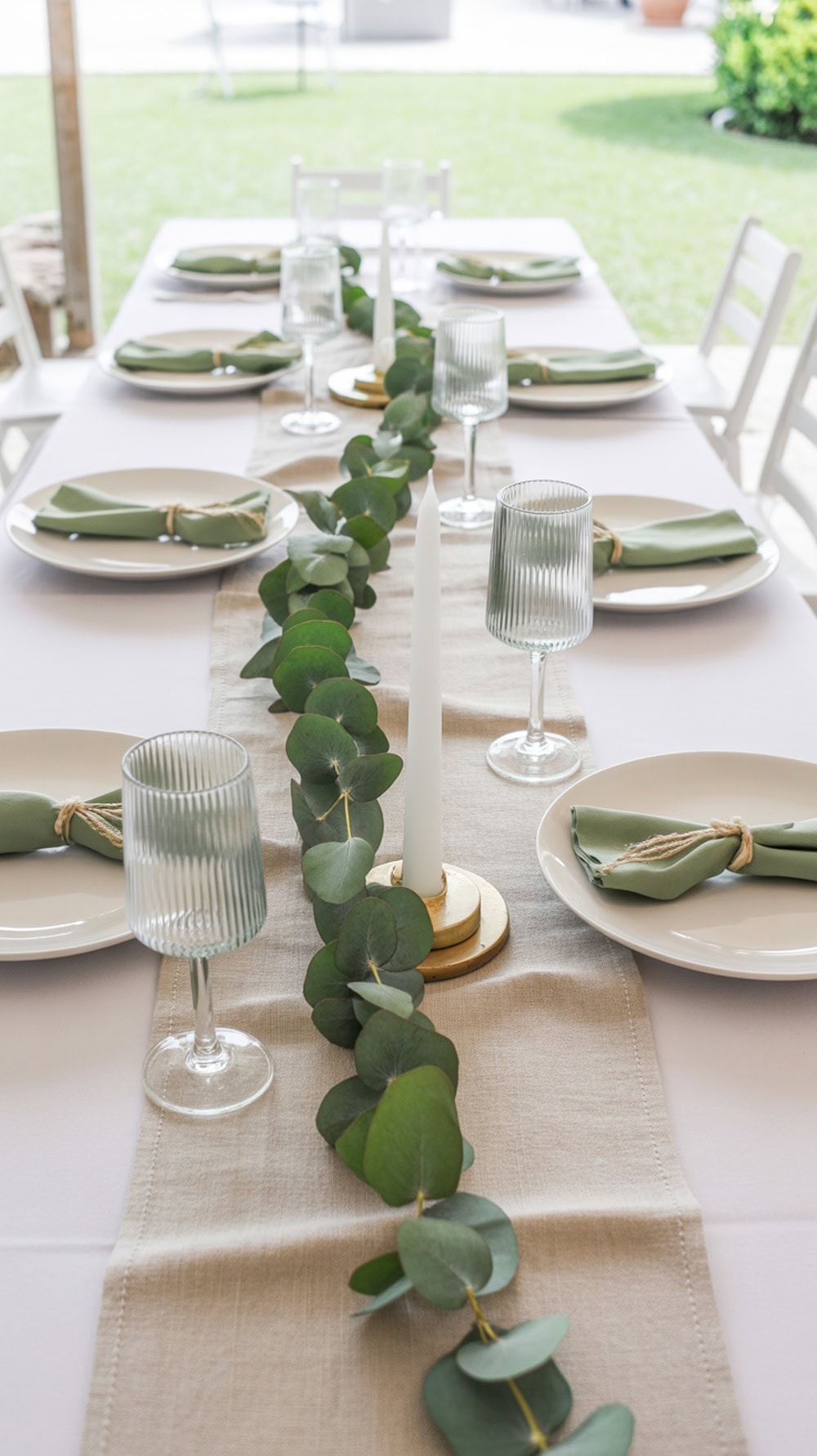 A modern minimalist graduation party table setup featuring white tablecloth, green eucalyptus garland, white plates, green napkins, and clear glassware.
