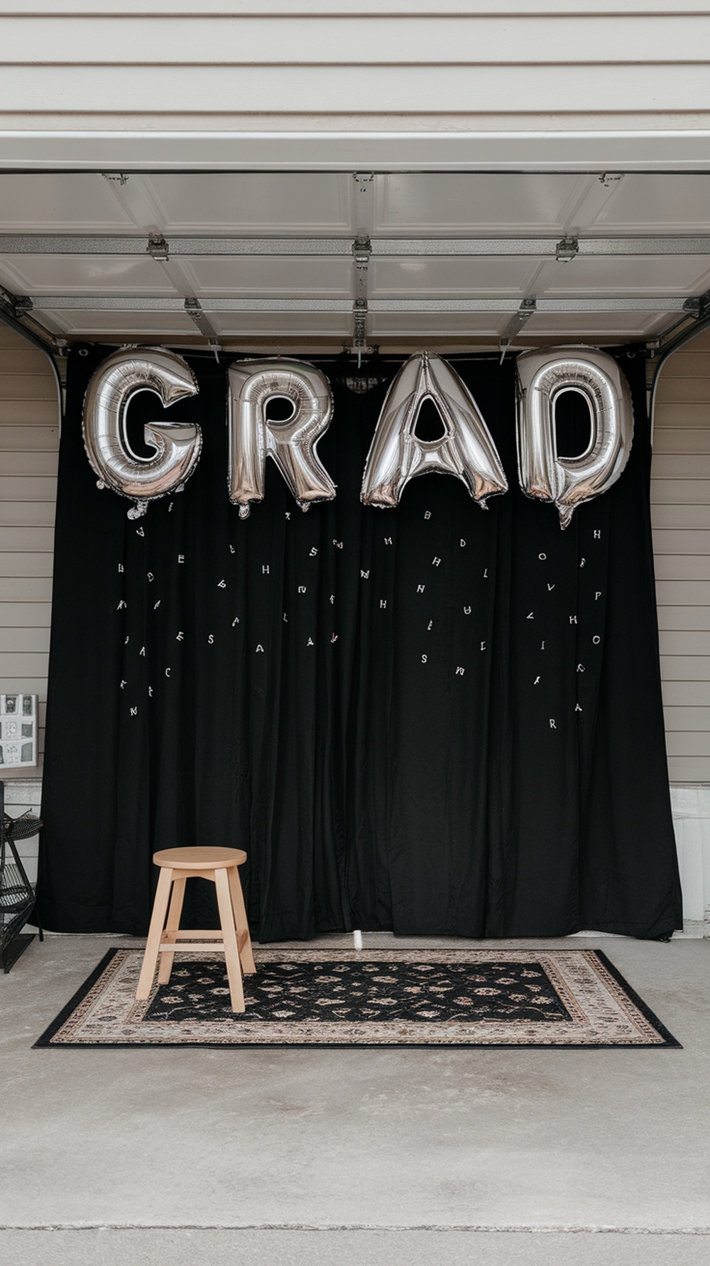 A stylish garage setup featuring a black curtain, silver balloons spelling 'GRAD', a stool, and a patterned rug.