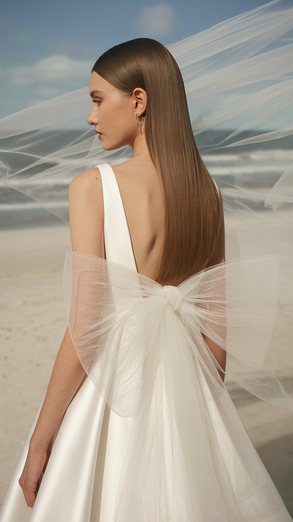 A bride with sleek straight hair styled with a statement veil bow, standing on a beach.