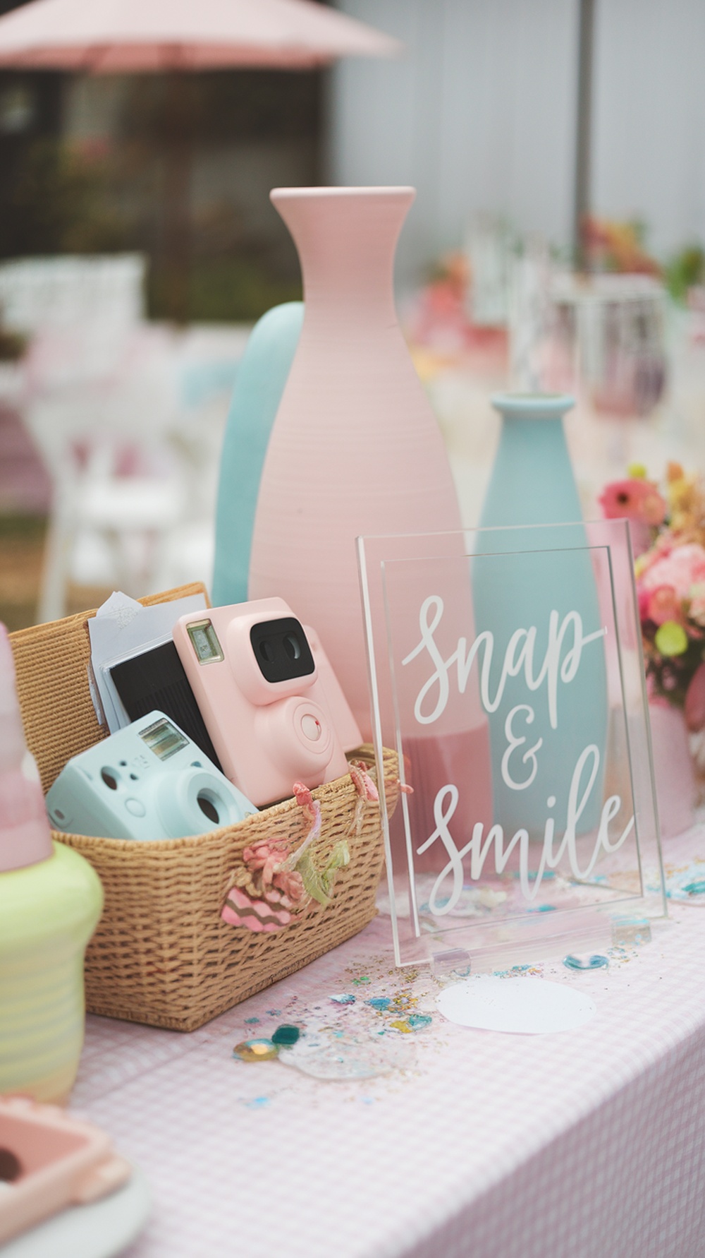 A pink-themed graduation party table with Polaroid cameras, pastel vases, and a sign that says 'Snap & Smile'.