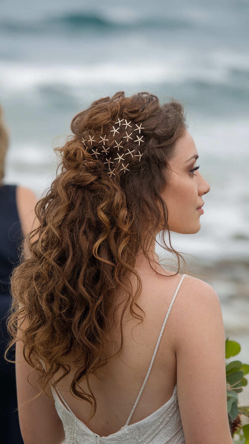A woman with curly hair styled in a half-updo, adorned with delicate starfish pins, standing by the beach.